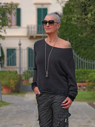 Ältere Frau trägt den OFF#DLY Pulli Sanja black: Ein schwarzer, locker fallender Off-Shoulder-Pullover mit leichtem Glanz und langen Ärmeln, kombiniert mit einer langen Halskette.