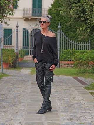 Die Frau trägt den OFF#DLY Pulli Sanja black, einen lässig-eleganten, off-shoulder Feinstrick-Pullover, kombiniert mit crinkle Cargo-Hosen und Stiefeletten, vor einem schmiedeeisernen Tor.