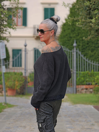 Ältere Frau trägt den OFF#DLY Pulli Sanja black mit lässigem Off-Shoulder-Design, enthüllt Tattoo; kombiniert mit dunklen Cargo-Hosen, stehend vor dekorativem Tor und Villa.