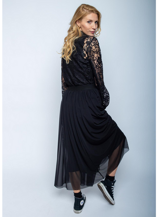 Junge Frau trägt den Emily Rock 201451 black, einen ausgestellten, semi-transparenten, knöchellangen Rock mit Gummibund und offener Saumkante, kombiniert mit einem schwarzen Spitzenoberteil und Converse-ähnlichen Sneakers.