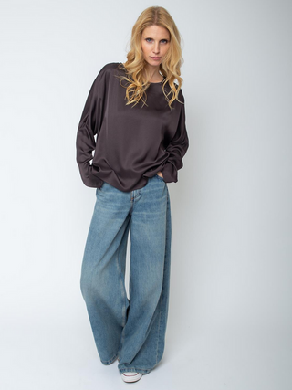 Person trägt Emily Bluse 157490-10 chocolate, eine locker sitzende, langärmelige Satinbluse mit rundem Ausschnitt und überschnittenen Schultern, kombiniert mit weit geschnittenen Jeans und weißen Sneakers.