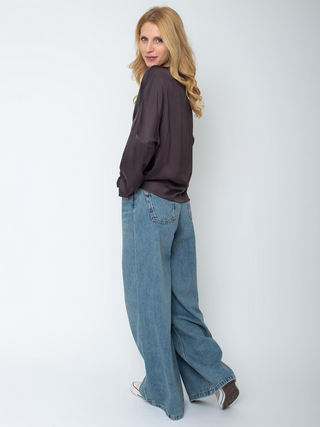 Die Emily Bluse 157490-10 chocolate, getragen von einer Frau, zeigt einen fließenden, satinierten Stoff mit rundem Ausschnitt und modernen, überschnittenen Schultern, kombiniert mit weit geschnittenen Jeans.
