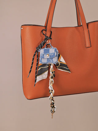 OFF#DLY Taschenanhänger Denimblue: Dekorativer Anhänger mit blau-weiß kariertem Stoffbeutel, schwarzem Kordelband, mehrfarbigem Seidenstück und geflochtener Kette mit goldfarbenem Clip an mittelgroßer cognacbrauner Tasche.