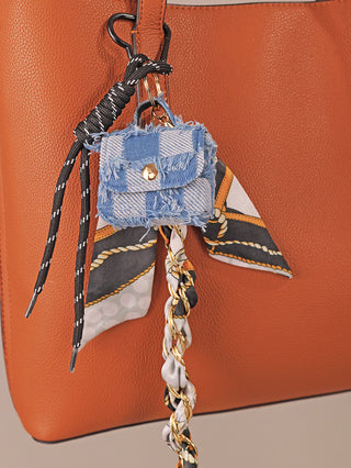 Nahaufnahme eines orange-braunen Ledertote mit dem OFF#DLY Taschenanhänger Denimblue, bestehend aus einem fransigen Denim-Check Minipouch, einem bedruckten Schal, einer schwarz-weißen Kordel und einer geflochtenen goldenen Kette.