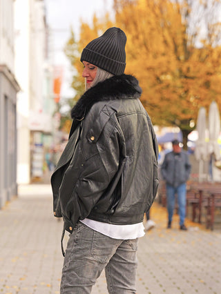 Eine Person trägt die OFF#DLY Mütze Anna black, eine schwarze Strickmütze, kombiniert mit einer schwarzen Lederjacke und grauen Jeans, während sie auf einem herbstlichen Bürgersteig steht.