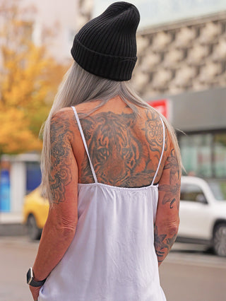 Person von hinten in urbaner Umgebung trägt ein OFF#DLY Top Button white, ein locker fallendes, weißes Camisole-Top mit dünnen Trägern und Knopfleiste, kombiniert mit schwarzer Strickmütze.