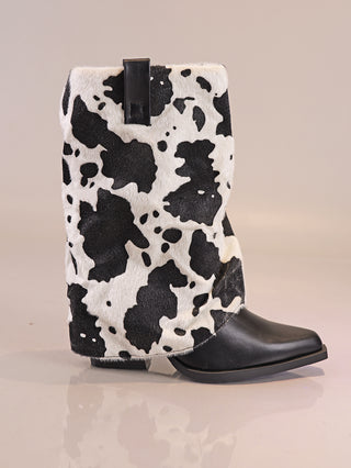 OFF#DLY Cow Boot black/white, ein Mid-Calf-Stiefel mit schwarzem Lederfuß und schwarzem-weißen Kuhfellmuster-Schaft, steht auf einer glatten Oberfläche. Oberer Schaft mit kleinem, schwarzem Zugriemen.