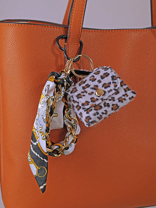 Nahaufnahme des OFF#DLY Taschenanhänger Leo lightblue, bestehend aus einem Leopardenmuster-Mini-Geldbeutel und einer goldenen Kette mit bedrucktem Schal, dekorativ an einem tanfarbenen Leder-Totebag befestigt.