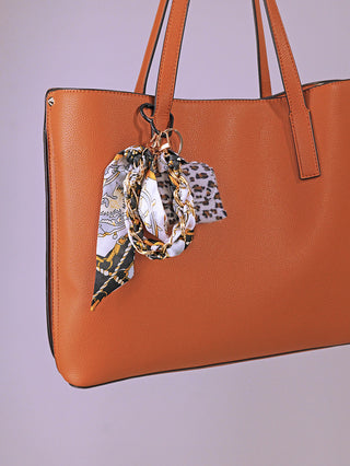 Nahaufnahme eines strukturierten Taschenanhängers OFF#DLY Leo lightblue, bestehend aus einem bedruckten Seidentuch, einer goldfarbenen Kette und einem Leopardenmuster-Stoffstück, an einer cognacfarbenen Ledertasche befestigt.