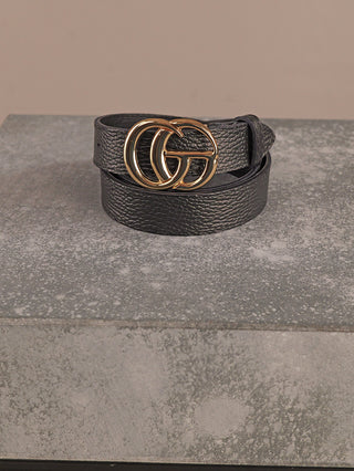 Der OFF#DLY Ledergürtel CG black zeigt einen schwarzen, genarbten Ledergürtel mit einer glänzenden, goldfarbenen Doppel-G-Schnalle, stilvoll auf einer grauen, strukturierten Steinoberfläche präsentiert.