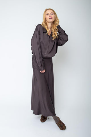 Eine blonde Frau trägt die Emily Bluse 155923-10 chocolate, eine locker sitzende, langärmelige Bluse mit Knopfblende, kombiniert mit einem passenden, bodenlangen Rock und braunen Mokassin-Slippern.