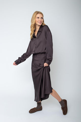 Emily Bluse 155923-10 chocolate: Frau in lockerem, dunklem Seidenhemd mit Knopfleiste und passendem Midirock, kombiniert mit grauen Socken und braunen Mokassins, vor weißem Hintergrund in leichter Bewegung.