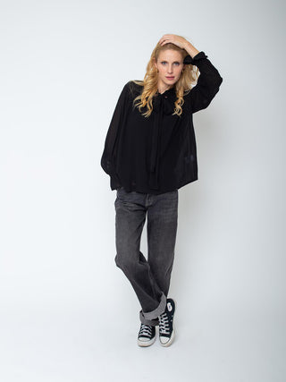 Frau trägt die Emily Bluse 157032 black mit Schluppe und Raglanärmeln, kombiniert mit geraden Jeans und schwarzen Sneakers, posiert lässig mit Hand am Kopf und gekreuzten Beinen.