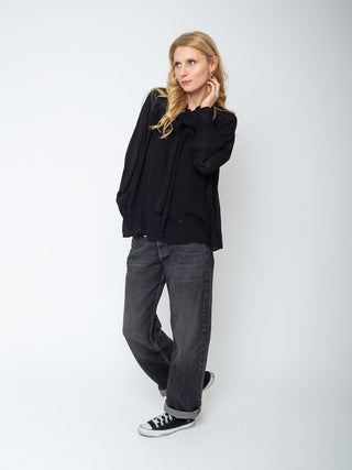 Emily Bluse 157032 black: Locker geschnittene, schwarze Bluse mit Schluppe, leicht ausgestellten Raglanärmeln und Smok-Details am Ärmelsaum, getragen von einer Frau in einer hellen Studioaufnahme.