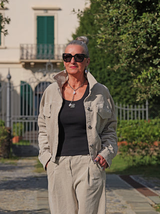 Frau in OFF#DLY Jacke Vera sand mit hochgestelltem Kragen, kombiniert mit passender Hose und schwarzem Top, steht lässig in einer Villaeinfahrt. Accessoires: Sonnenbrille, Kette, Ohrringe.