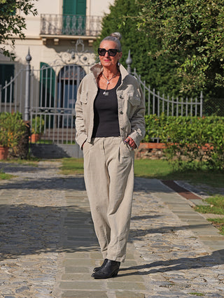 Frau trägt die OFF#DLY Jacke Vera sand mit lässiger Cord-Optik, lockerem Schnitt und Knopfleiste, während sie auf einem gepflasterten Weg durch einen Garten geht. Accessoires ergänzen den stilvollen Look.