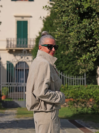 Person trägt die OFF#DLY Jacke Vera sand, eine lässige, kastige Cordjacke mit Kragen und Knopfleiste, ideal für Layering. Sie steht vor einem Gebäude mit grünen Fensterläden.