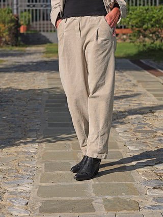 Person trägt OFF#DLY Pant Vada sand, beige Cordhose mit weitem Schnitt und umgeschlagenem Saum, kombiniert mit schwarzen, spitz zulaufenden Stiefeletten auf einem Steinweg.