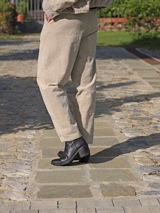 OFF#DLY Pant Vada sand: Person trägt beige Cordhose mit geradem Schnitt und Umschlag am Saum, kombiniert mit schwarzen Schnürstiefeln, stehend auf unebenem Steinweg.