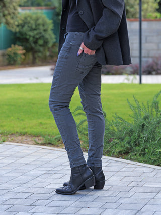 Person trägt OFF#DLY Color Jeans Imelda grau, slim-fit, in schwarzen Lederstiefeln. Hand in der Tasche, kombiniert mit dunkler Jacke. Ideal für stilvolle Alltags-Looks.