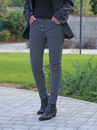 Person trägt OFF#DLY Color Jeans Imelda grau, schlanke Passform mit Knopfleiste, kombiniert mit einem dunklen Blazer und schwarzen Lederstiefeln, Hände in Taschen, teilweise sichtbare Ringe und dunkler Nagellack.