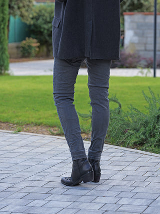 Person trägt OFF#DLY Color Jeans Imelda grau, schlanke Passform. Kombiniert mit schwarzen Lederstiefeletten und dunklem Mantel. Steht auf gepflastertem Weg, umgeben von gepflegtem Garten.