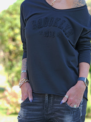 Person trägt das OFF#DLY LS Brooklyn black: schwarzes, schulterfreies Langarmshirt mit „Brooklyn NYC“-Stickerei, kombiniert mit Jeans, Armbändern, Ringen, Smartwatch, Kette und sichtbarem Tattoo am Unterarm.