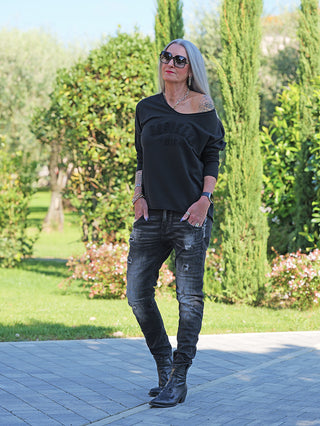 Ältere Frau trägt OFF#DLY LS Brooklyn black Longsleeve, oversized, off-shoulder, mit „Brooklyn NYC“-Stickerei. Kombiniert mit dunklen, leicht distressed Jeans und schwarzen Stiefeletten im Garten.