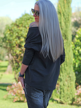 Person trägt das OFF#DLY LS Brooklyn black Longsleeve, mit weitem Ausschnitt und „Brooklyn NYC“-Stickerei, im Garten. Sichtbar sind lange, silberne Haare und ein Tattoo auf der Schulter.