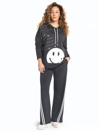 Frau trägt Doris Streich Shirt 327150 schwarz/weiß, ein schwarzes Sweatshirt mit großem Smiley auf der Vorderseite, weißen Kordeln, kombiniert mit lockeren schwarzen Hosen mit weißen Streifen und weißen Sneakers.