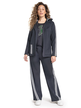 Frau in schwarzem Doris Streich Relax Hose schwarz/weiß Athleisure-Set mit weißen Streifen, bestehend aus Kapuzenjacke, T-Shirt mit grünem Grafikmotiv und passenden Trackpants, posiert lässig, weiße Slip-ons tragend.