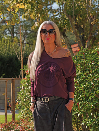 Person trägt das OFF#DLY LS Vintage bordeaux: ein lässiges, off-shoulder Longsleeve mit geprägtem Emblem, kombiniert mit weit geschnittenen Karohosen und auffälligen Accessoires im Garten.