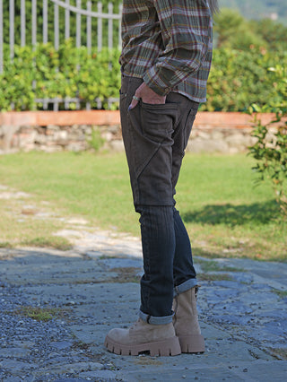 Person trägt OFF#DLY Sina Brown Wash Denim, dunkle slim-fit Jeans mit hochgekrempelten Bündchen und markanten Nähten, kombiniert mit robusten, beigen Wildlederstiefeln auf einem Steinweg.