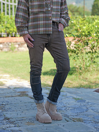 Person trägt OFF#DLY Sina Brown Wash Denim, schlanke Jeans mit Patch-Details und Cargo-Tasche. Kombiniert mit kariertem Flanellhemd und beigen Schnürstiefeln, betont durch Schmuck und lackierte Fingernägel.