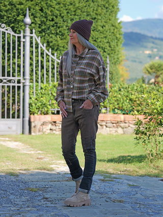 Frau in OFF#DLY Sina Brown Wash Denim mit Nahtdetails und Used-Look, kombiniert mit karierter Bluse, Strickmütze und robusten Boots, stehend auf Steinpfad vor einem Gartentor.