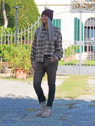 Person trägt die OFF#DLY Bluse Amelia multicolor, eine locker geschnittene, karierte Bluse mit Knopfleiste und Brusttaschen, kombiniert mit Jeans und Stiefeln, vor einem dekorativen Tor.