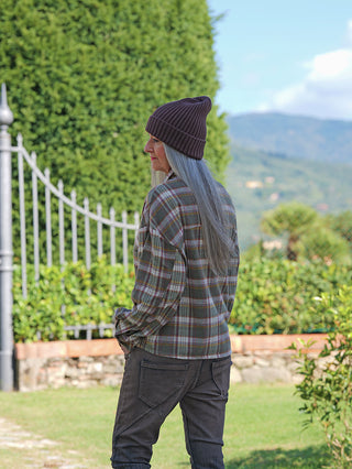 Person von hinten in einem Garten, trägt die OFF#DLY Bluse Amelia multicolor mit Karomuster und langen Ärmeln, kombiniert mit einer dunklen Hose und einer braunen Mütze.