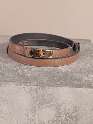 OFF#DLY Gürtel Leder taupe, ein schlanker Ledergürtel mit goldfarbener Drehverschluss-Schnalle, liegt gerollt auf einem Betonblock. Die Rückseite ist schwarz, Breite 2 cm, größenverstellbar.