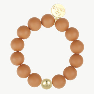 Elastisches Kugelarmand caramel 64: Ein stretch‐Stil Armband mit matten, runden, tanfarbenen Perlen und einer glänzenden Goldperle. Ein goldfarbener Anhänger zeigt GINUO und ein Logo.