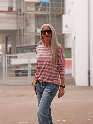 Frau trägt ein OFF#DLY Ringel Dora taupe/rosa Shirt mit weiten Streifen, kombiniert mit blauer Jeans und Schmuck, vor urbanem Hintergrund.