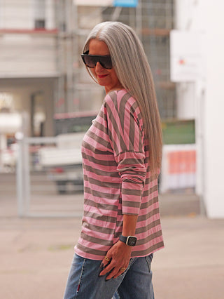 Person trägt das OFF#DLY Ringel Dora taupe/rosa, ein locker sitzendes, langärmeliges Shirt mit Rundhals, horizontalen Streifen in Rosa und Taupe, kombiniert mit blauer Jeans, im urbanen Hintergrund.