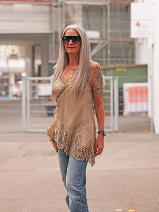 Frau trägt das OFF#DLY Top Violetta camel mit Spitzenbesatz. Das locker fallende Top harmoniert mit blauen Jeans und Accessoires, ideal für elegante und lässige Looks.