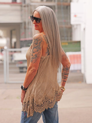 Frau trägt das OFF#DLY Top Violetta camel mit Spitzenabschluss, kombiniert mit Jeans und auffälligen Tattoos, Accessoires wie goldenen Armbändern und schwarzer Smartwatch, stilvoll und lässig präsentiert.