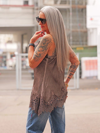 Person trägt das OFF#DLY Top Violetta braun, ein ärmelloses, locker geschnittenes Spitzentop, kombiniert mit blauen Jeans. Sichtbare Details: Spitze an Saum und Dekolleté, Tattoos, Accessoires.