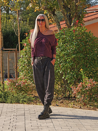 Frau trägt das OFF#DLY LS Vintage bordeaux Langarmshirt mit asymmetrischem Ausschnitt und Vintage-Emblem, kombiniert mit grauen Karohosen und schwarzen Kampfstiefeln, in einem lässigen, stilvollen Look.