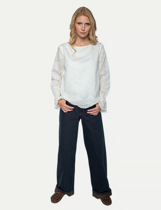 Die Emily Bluse 157110-10 offwhite/lace zeigt sich mit langem Ärmel und zarter Blumenstickerei, kombiniert mit einer wellenförmigen Saumkante. Sie wird von weiten, dunkelblauen Hosen ergänzt.