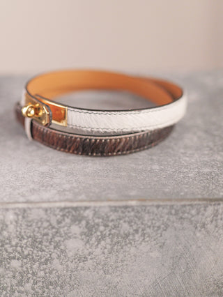 Zwei schlanke Ledergürtel, darunter der OFF#DLY Ledergürtel Fell braun, einer weiß mit Fellstruktur, der andere dunkelbraun, liegen aufgerollt auf einer grauen Oberfläche, beide mit goldfarbener Schließe.