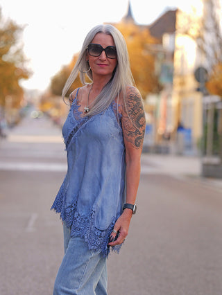 Frau trägt das OFF#DLY Top Violetta lightblue mit Spitzenverzierung am Ausschnitt und Saum, kombiniert mit hellen Jeans und Schmuck, während sie in einer herbstlichen Straße steht.