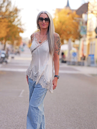 Person trägt das OFF#DLY Top Violetta champagner mit Spitzen-Details am Saum und Dekolleté, kombiniert mit hellblauer Jeans und Accessoires, in herbstlicher Straße.