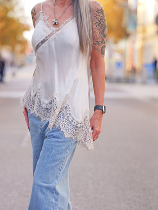 Person in lockerem, ärmellosem OFF#DLY Top Violetta champagner mit Spitzendetails, kombiniert mit hellblauen Jeans und einer dunklen Smartwatch am Handgelenk.